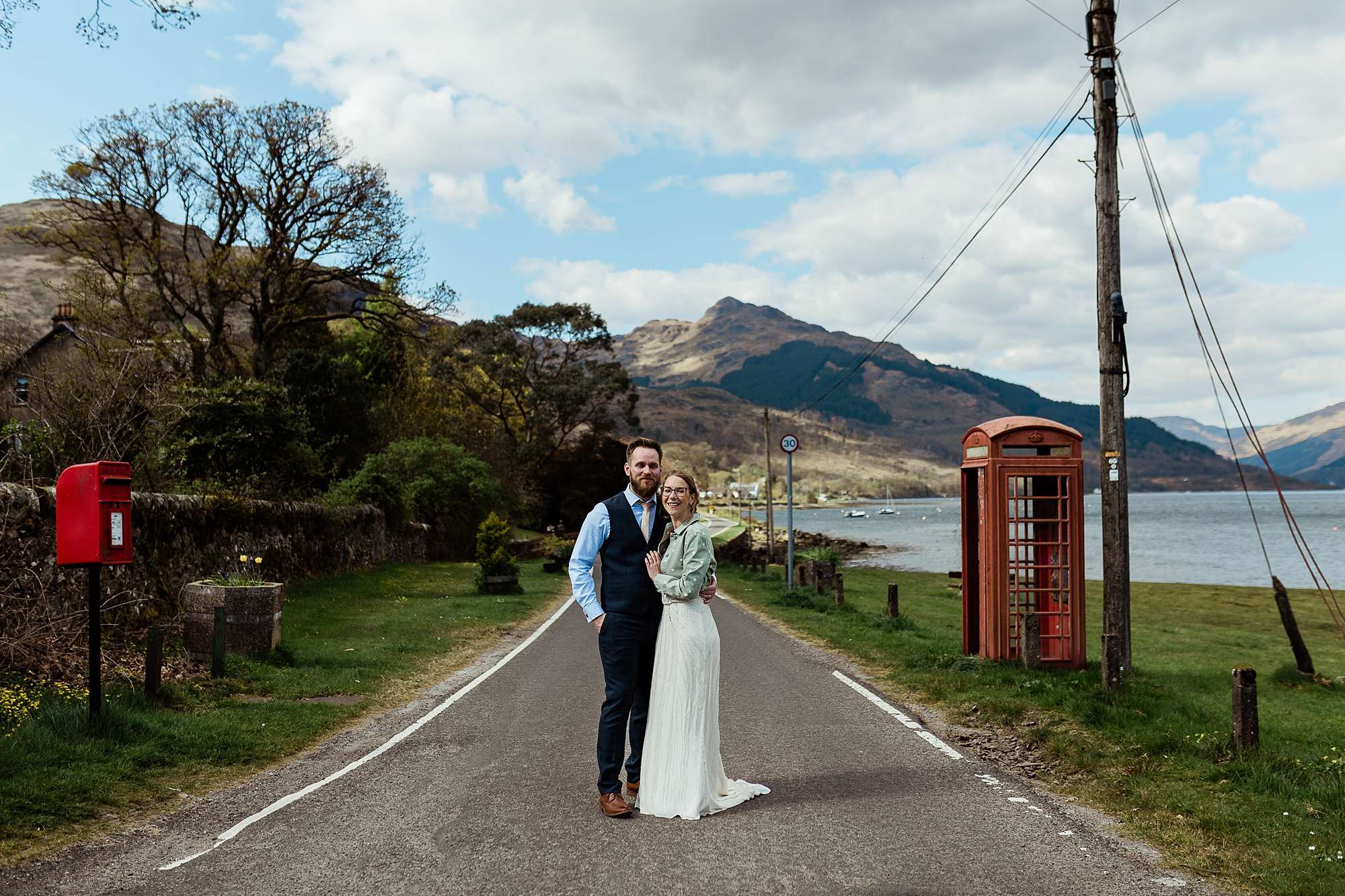 bride and groom standing on a scottish country road by a red post box and a red telephone box