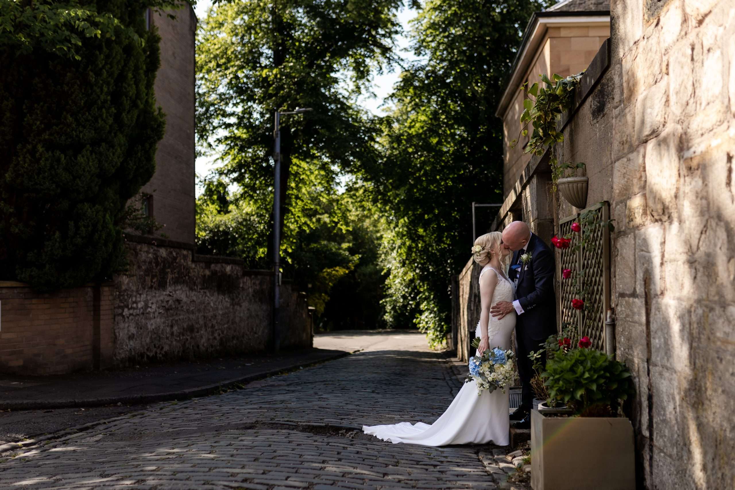 bride and groom kissing in a cobbled lane with trees and flowers next to One Devonshire Gardens