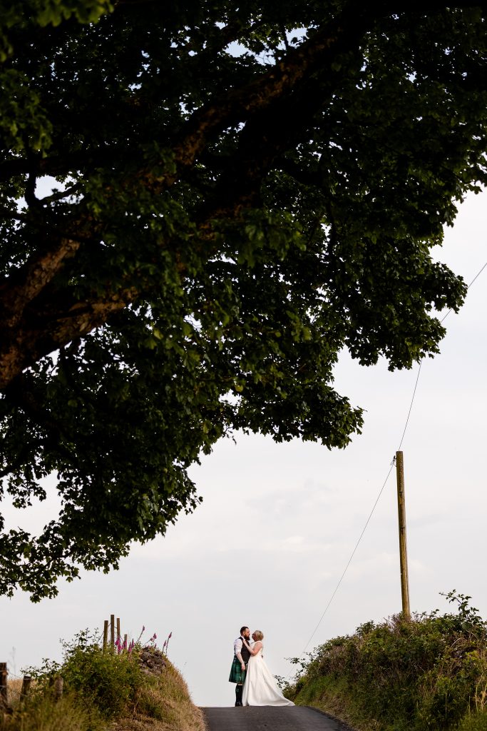 bride and groom standing on the crest of a hill on a country road under a tree