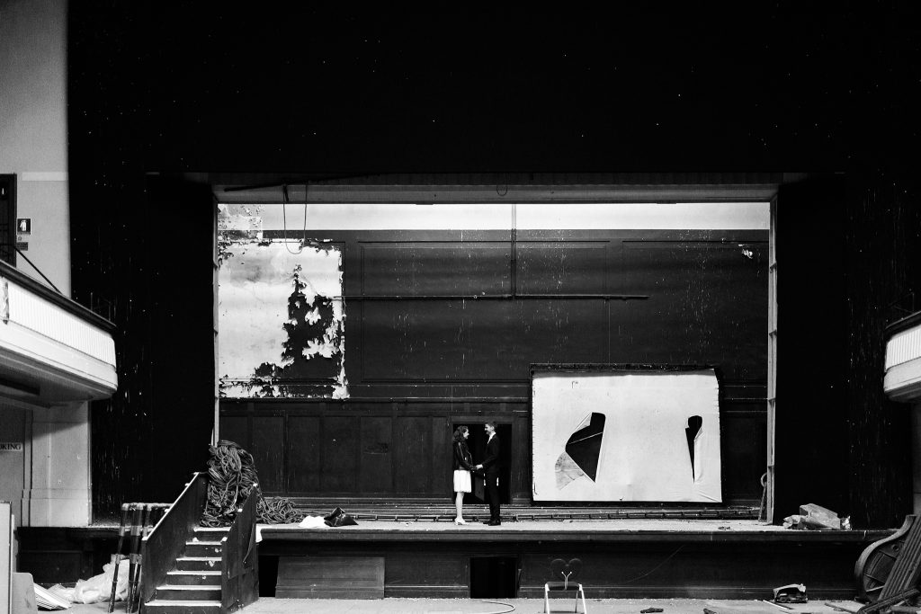 bride in a short white dress and black jacket, groom in a black suit, standing on stage in Leith Theatre at their wedding