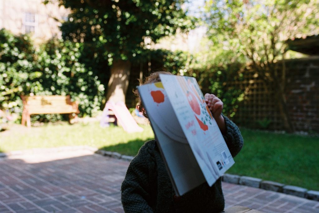 35mm photo of a child holding a book in front of their face in a garden during a during a documentary family photography session in glasgow