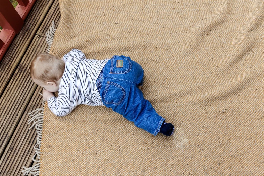 baby in blue jeans and a white stripey top crawling on a soft yellow blanket on decking in a garden 
