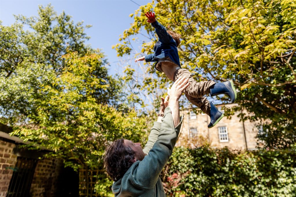 father throwing his little boy into the air outside in a garden in West End of Glasgow during a family photography session