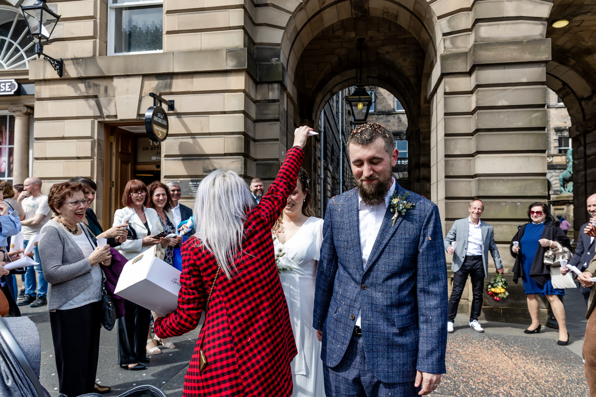 Guest in red houndstooth coat pouring confetti over newlyweds on Royal Mile next to City Chambers in Edinburgh