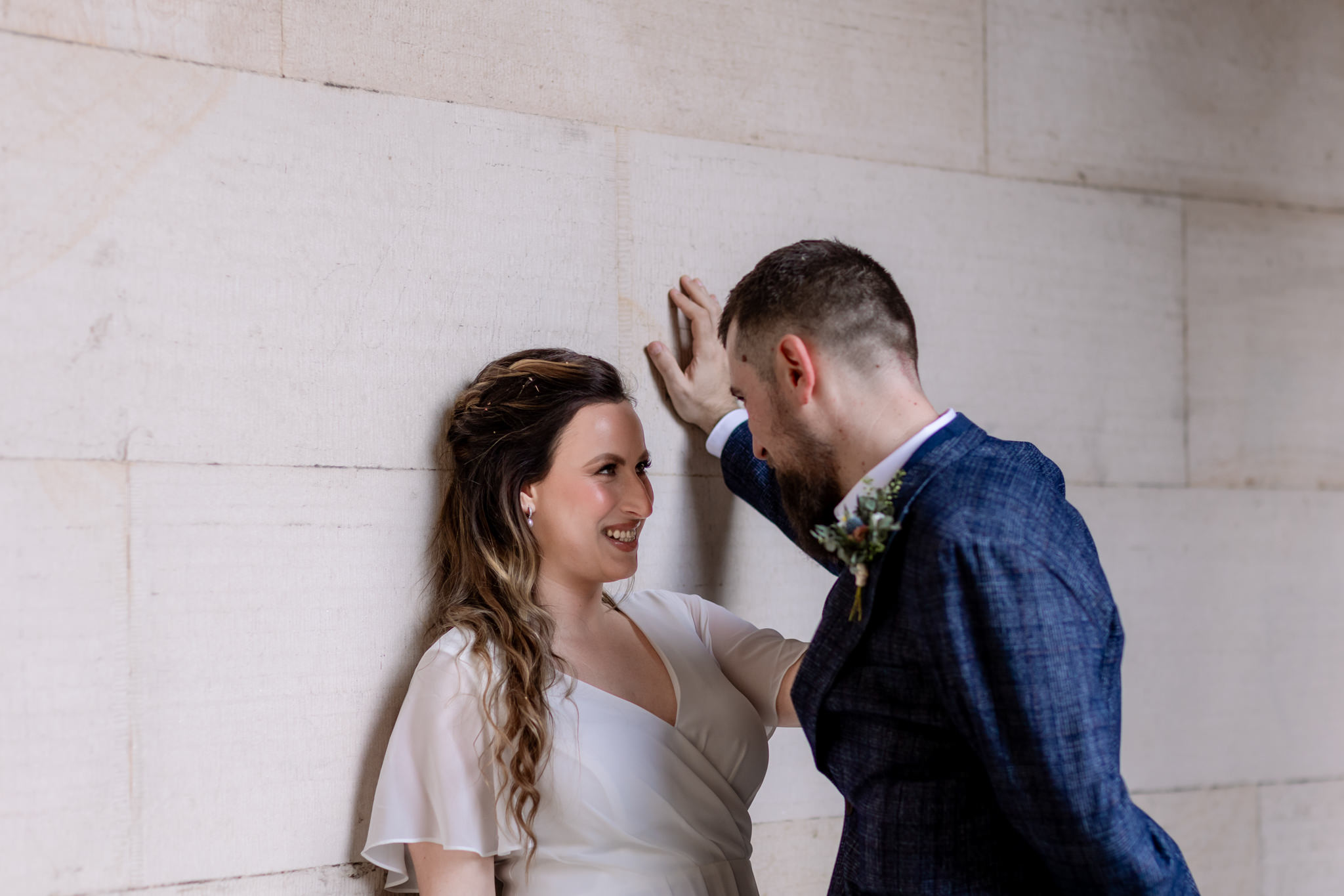 groom and bride smiling at each other against a soft standstone building