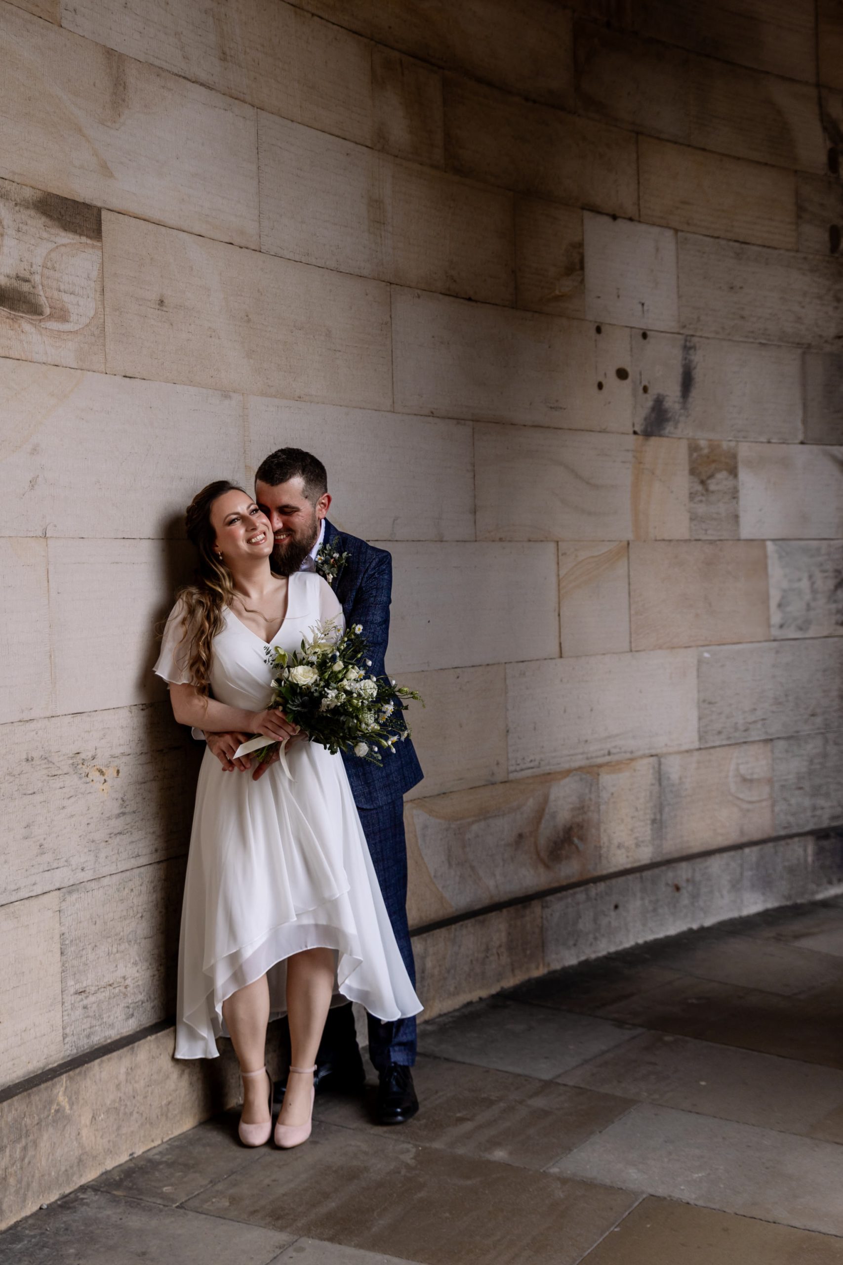 bride and groom embracing and smiling against a soft standstone building