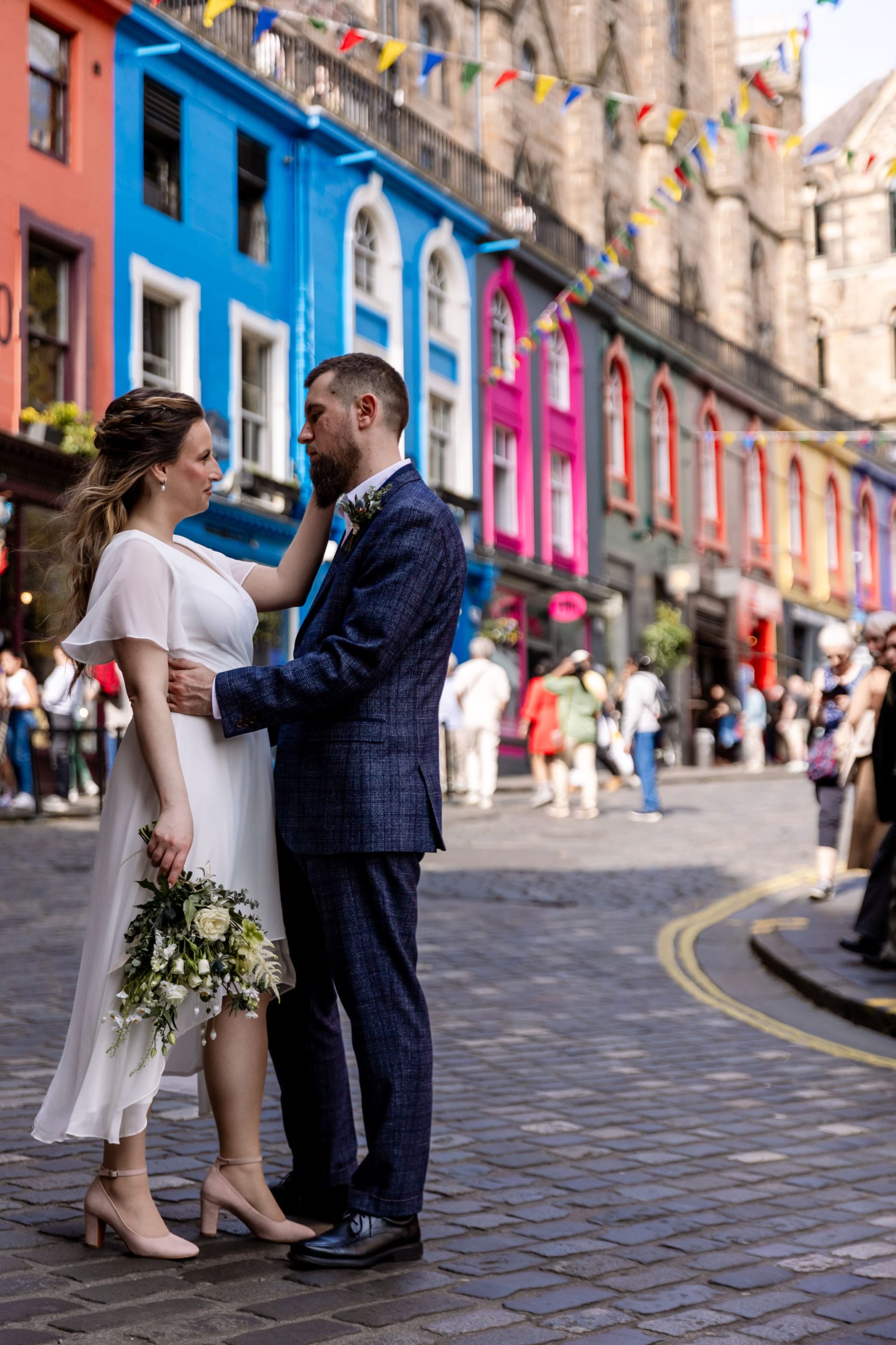 bride stroking groom's cheek at the bottom of Victoria Street in Edinburgh with bunting and multicolour shops 