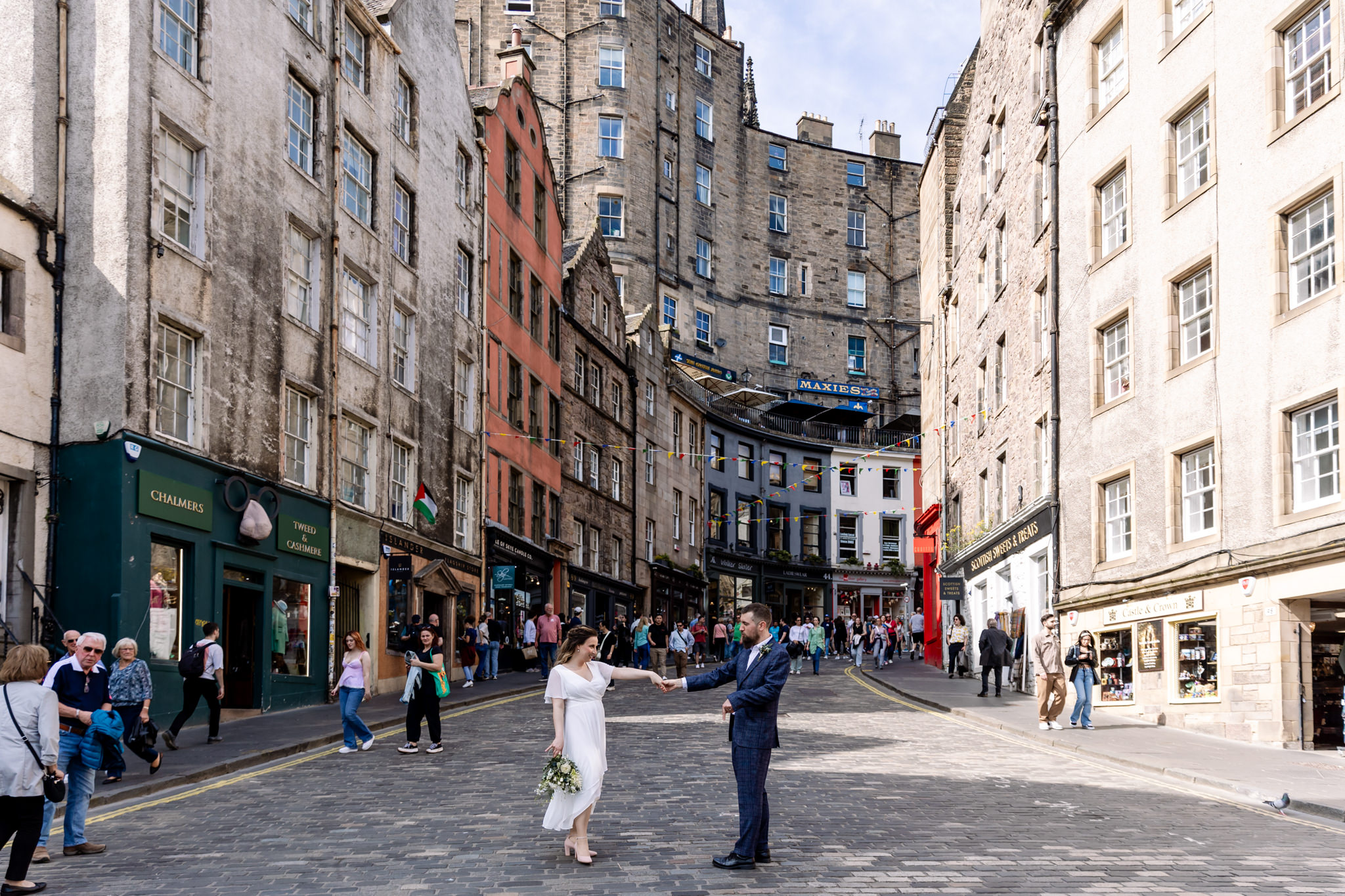 bride and groom dancing on cobble stones at foot of Victoria Street in Edinnburgh with bunting and multicolour shops 