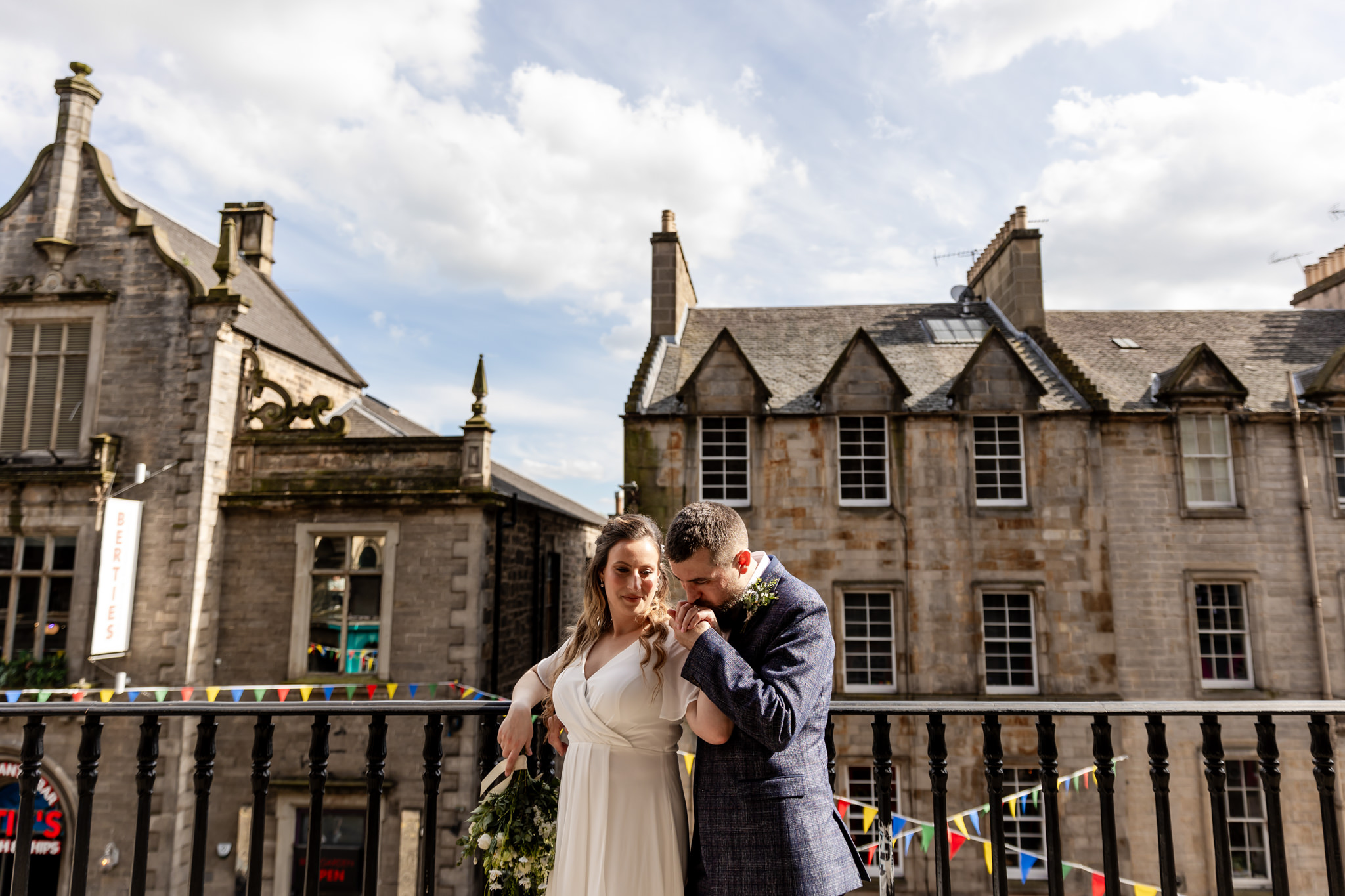 groom kissing bride's hand on Victoria Street terrace with buildings and bunting behind them 