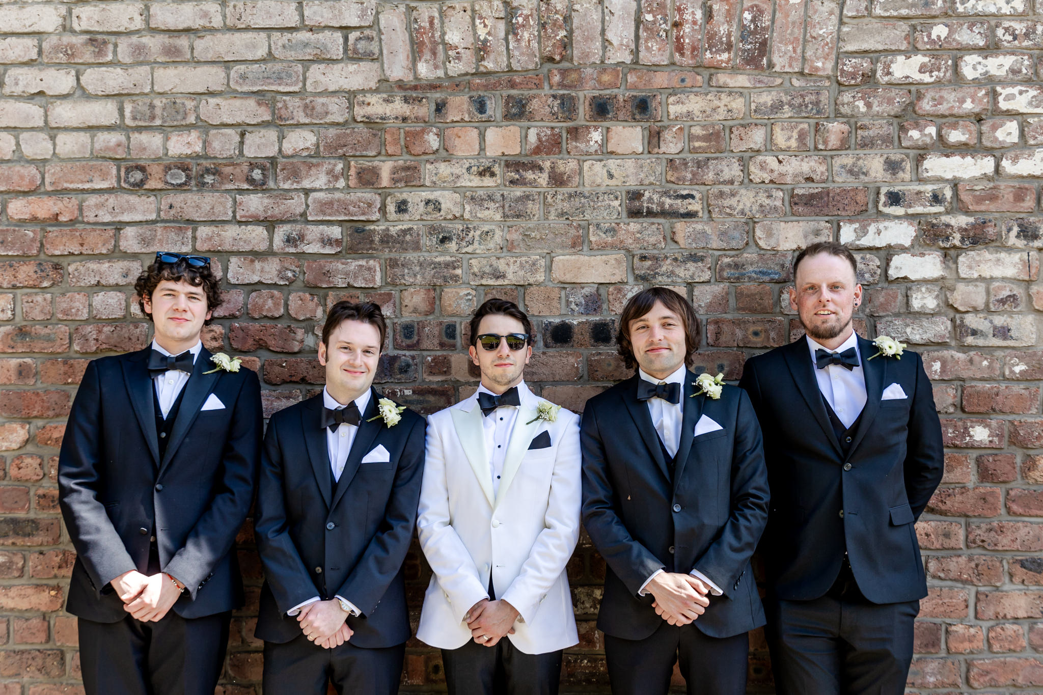 line up of four groomsmen in black tux and groom in a white tux against a brick wall 