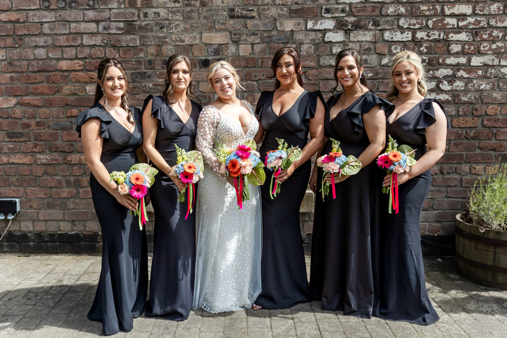 bride and bridesmaids in front of a brick wall. bridesmaids wear long black dresses, and all are holding bright floral bouquets