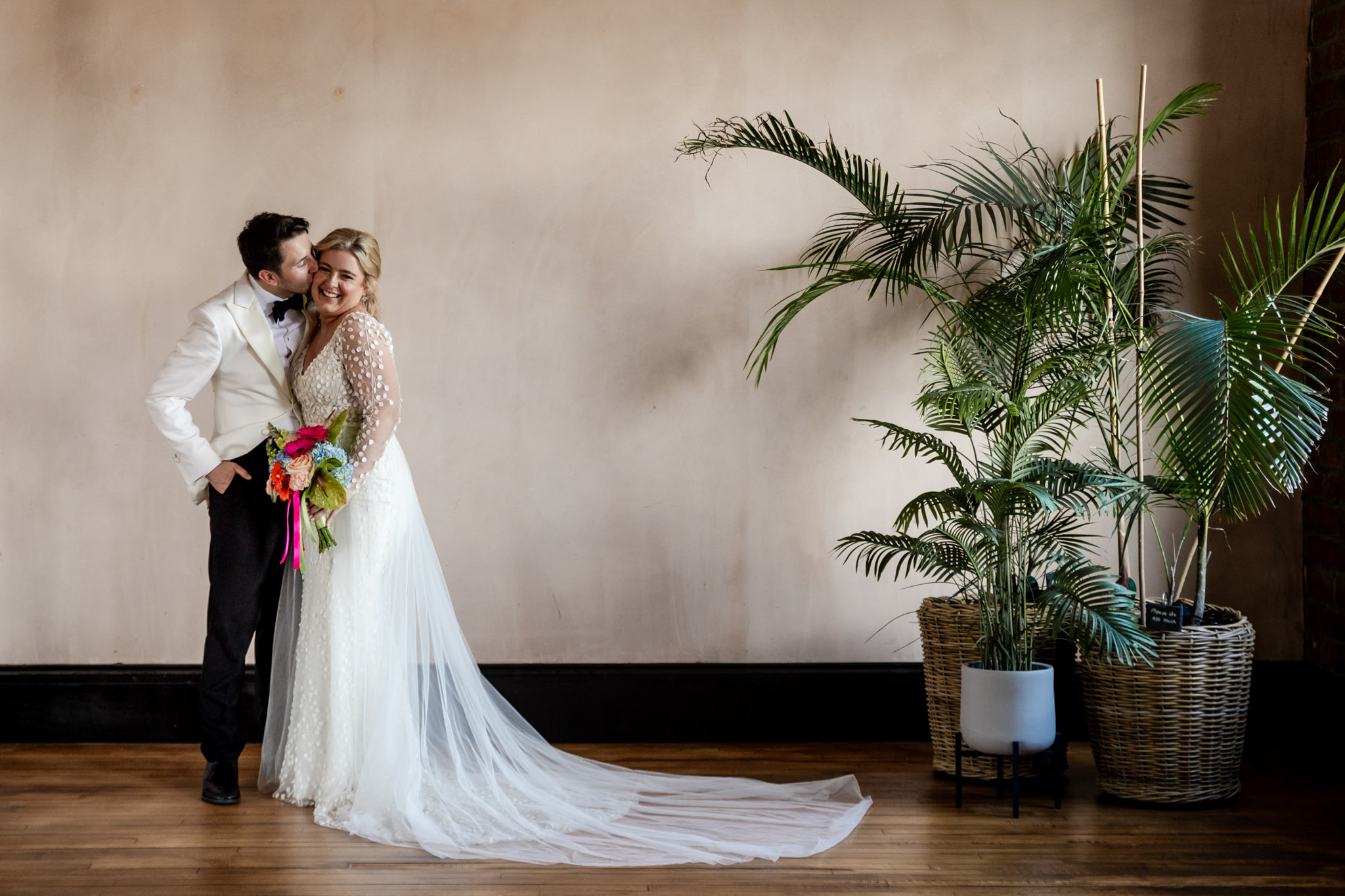 bride and groom kissing next to a peach coloured plaster wall and fern trees inside Engine Works