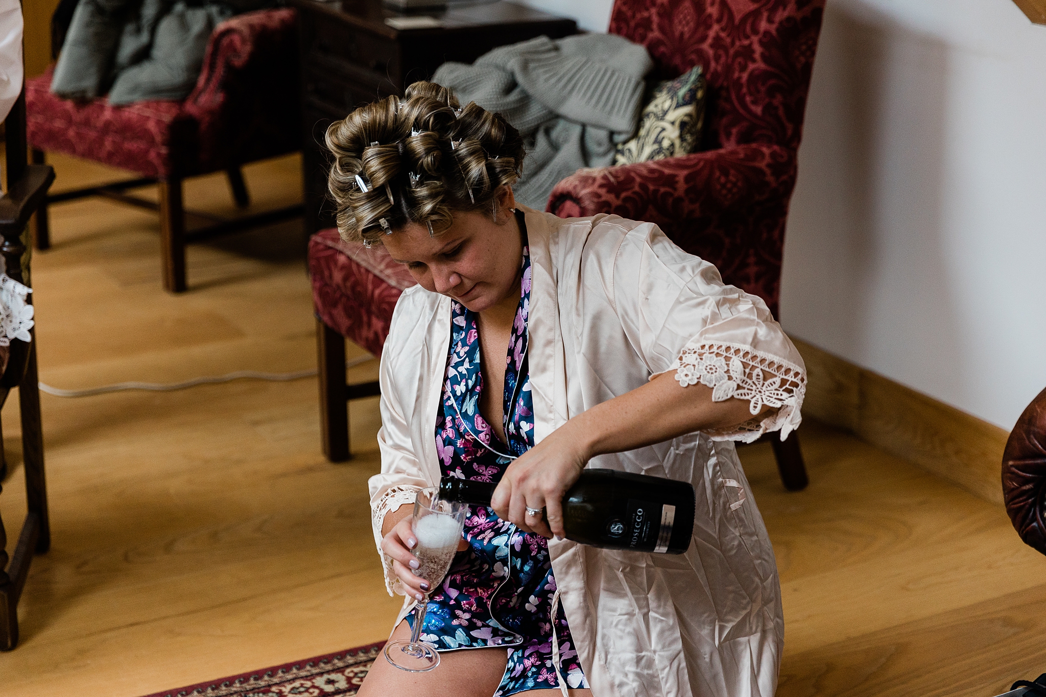 bridesmaid with hair in rollers and wearing a pink silk dressing gown pouring champagne into a glass