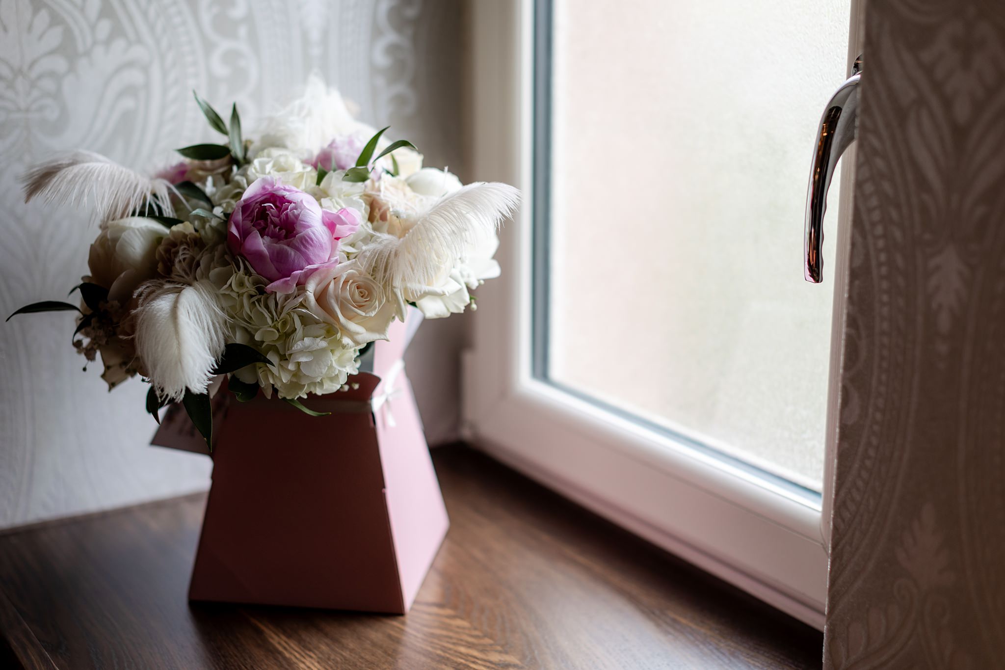 wedding bouquet with pink and white roses and feathers sitting in a box next to a window