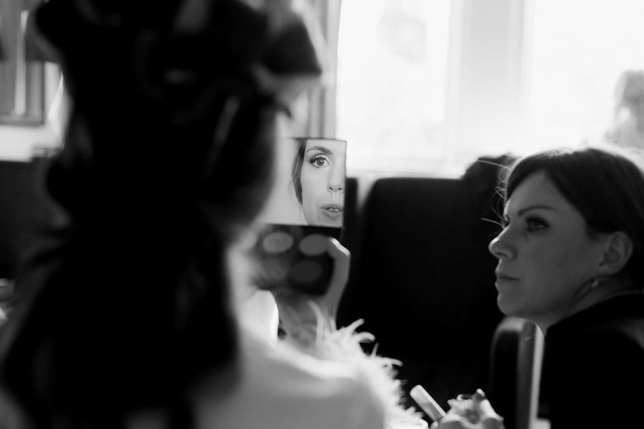 bride with her hair in rollers, wearing a pink pyjama top, checking her make up in a handheld mirror while her make up artist looks