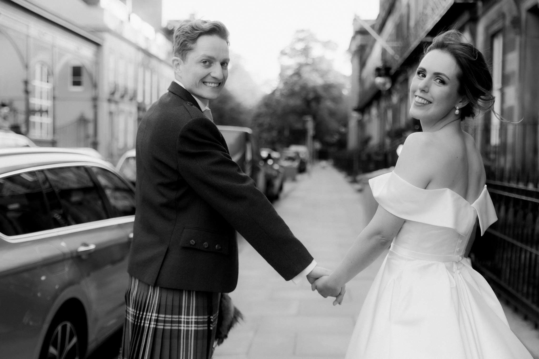 bride and groom walking down a street together looking back over their shoulder and smiling at the camera in Edinburgh 