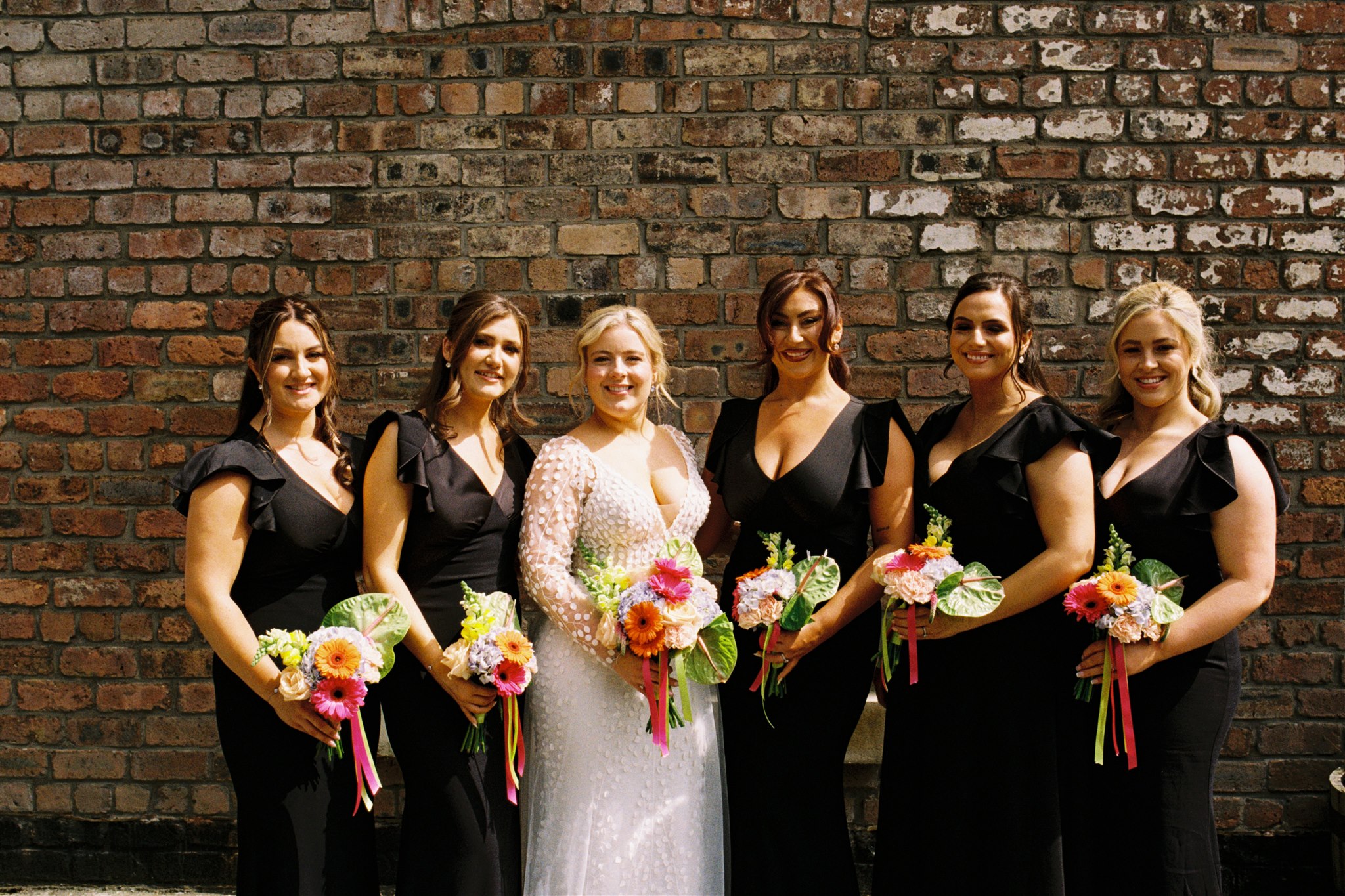 analogue film photograph of a bride and bridesmaids in black and white attire, standing outside a red brick wall in the sunshine 