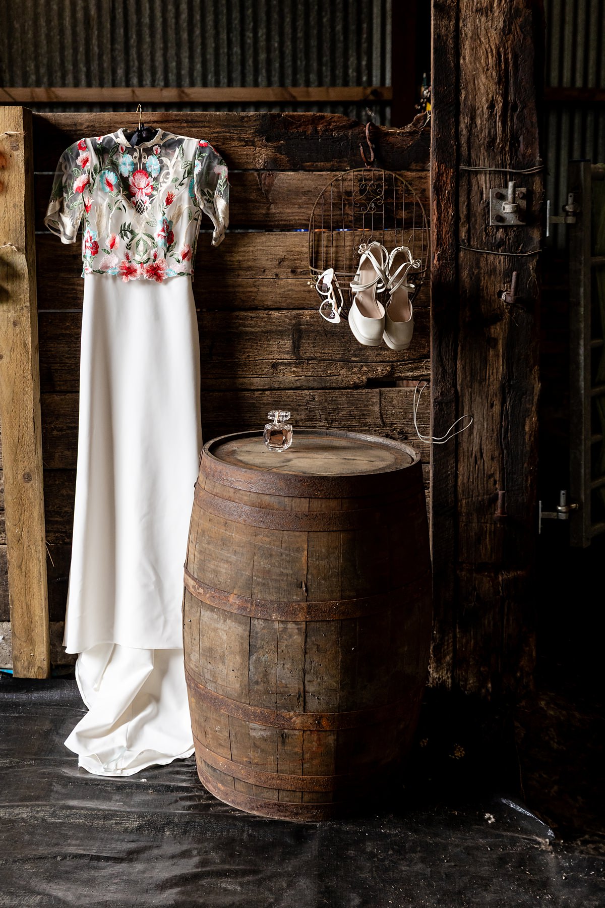 a bespoke wedding dress by Flossy and Dossy, hanging up in a barn. A bottle of perfume sits on a barrel in the foreground and a pair of white wedding shoes and sunglasses are hanging on a hanger.