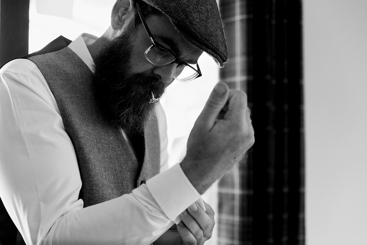 man attaching his cufflinks to his shirt. He's wearing a waistcoat, a flatcap, and has specs and a beard. He holds a cufflink in his mouth.