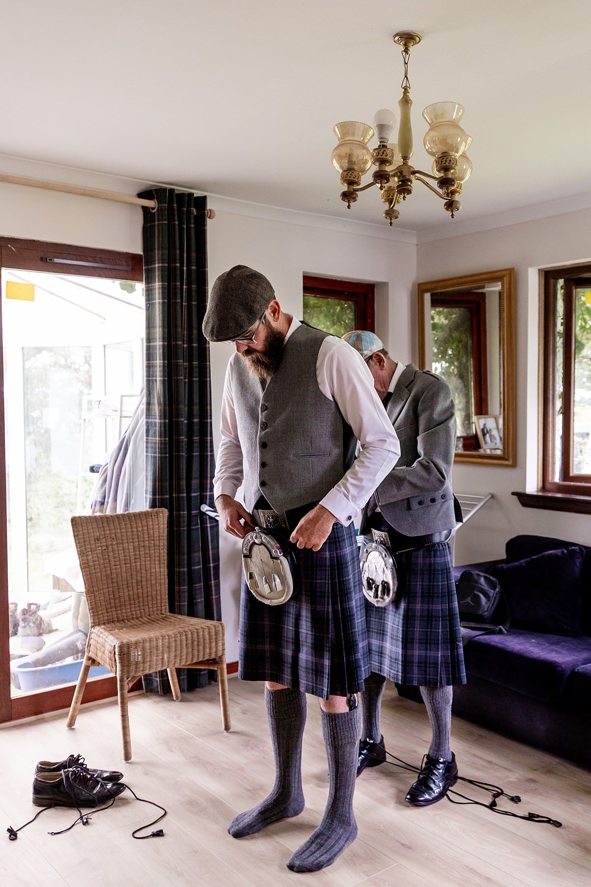an older man in a kilt helps a groom in a flat cap attach the sporan on his kilt.