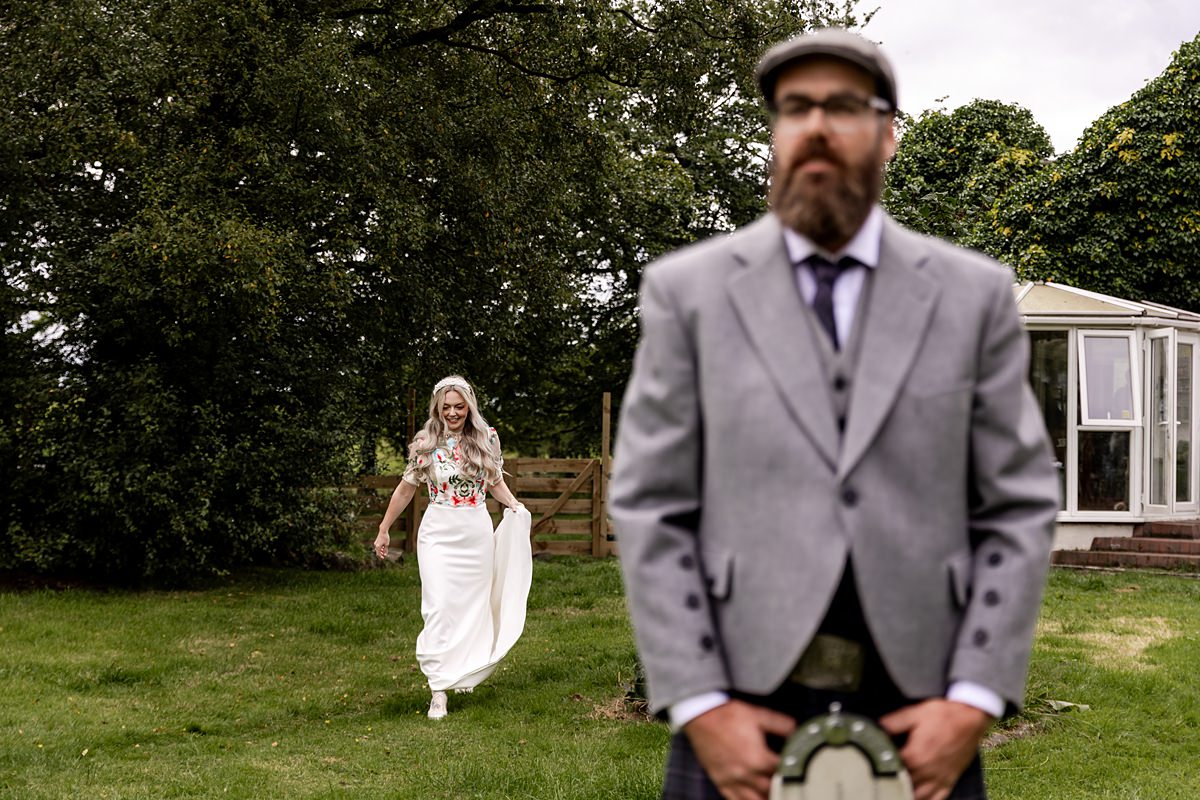 a man with a beard and glasses stands in the foreground wearing a kilt outfit and a flatcap. A woman in a wedding dress approaches him across a lawn from behind