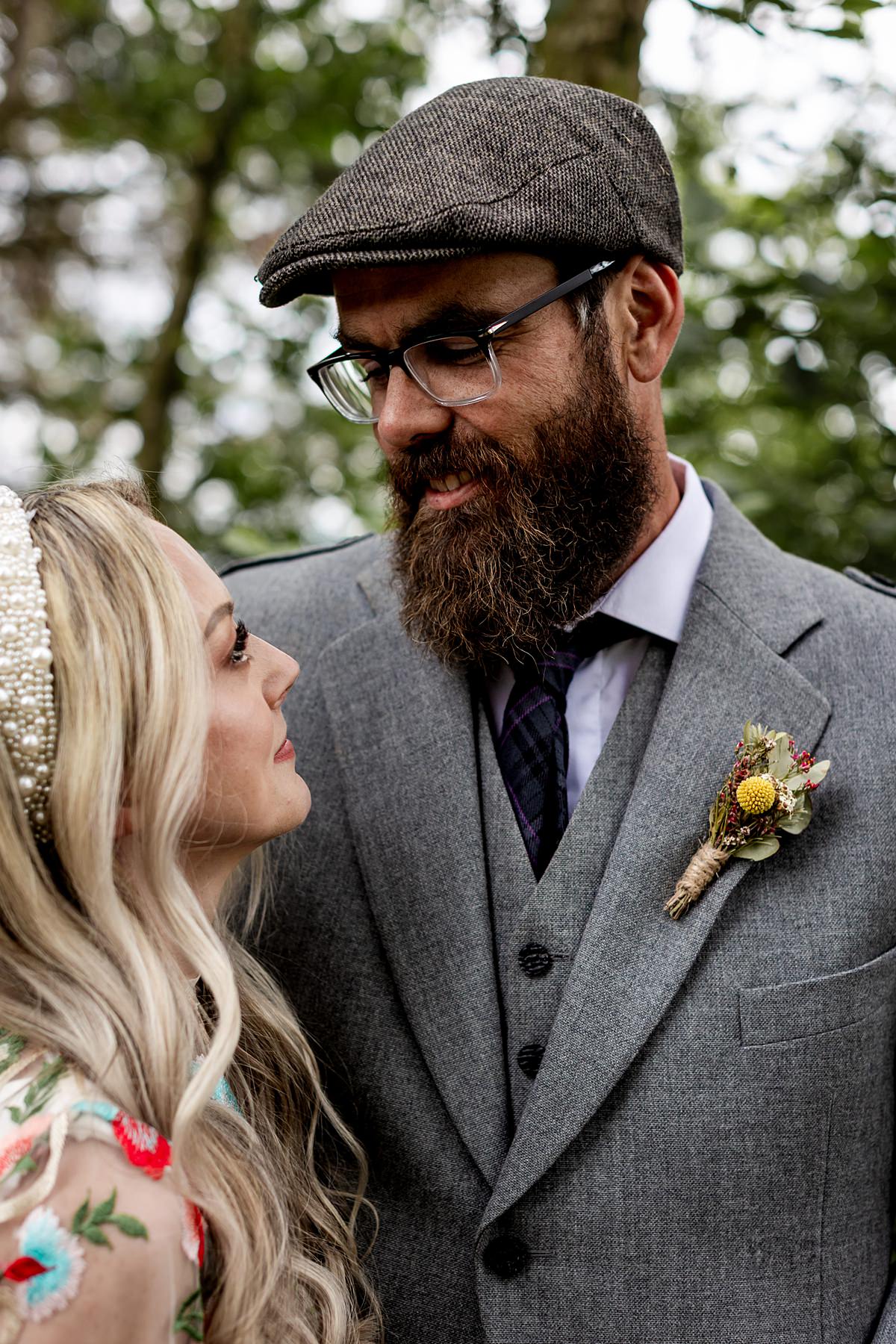 groom with specs and a beard and a flat cap looking at a blonde bride