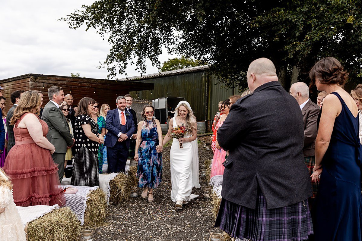 bride in a Flossy and Dossy dress, walking down an outdoor aisle, with her mother, surrounded by guests.