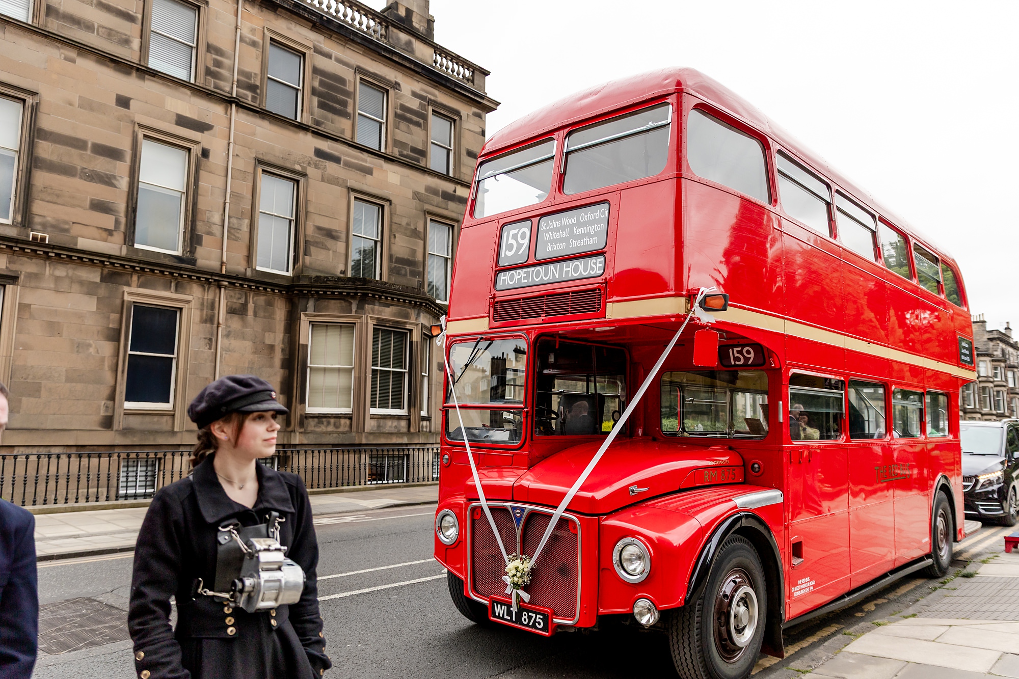 woman with a ticket machine standing next to a classic double decker red bus that reads Hopetoun House as the destination