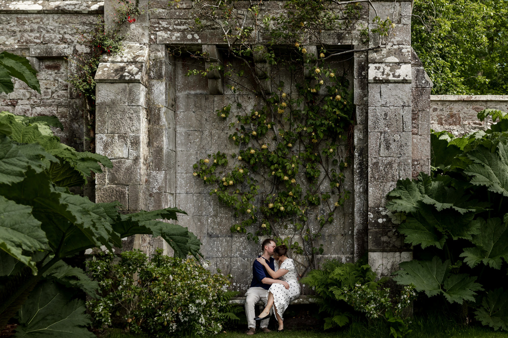 couple snuggling on a stone bench surrounded by foliage, in a castle garden