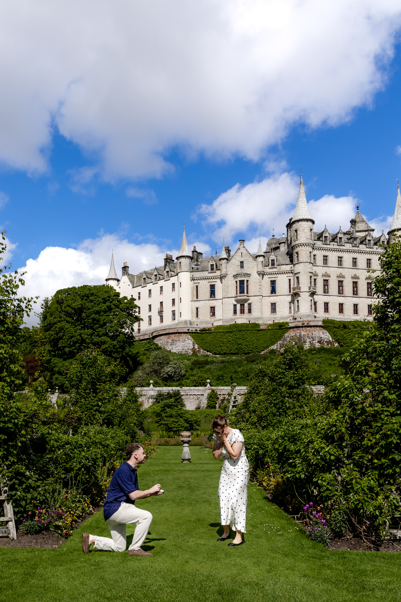 man kneeling to propose to his girlfriend in the garden with Dunrobin Castle in the background on a sunny day  in the Scottish Highlands. 