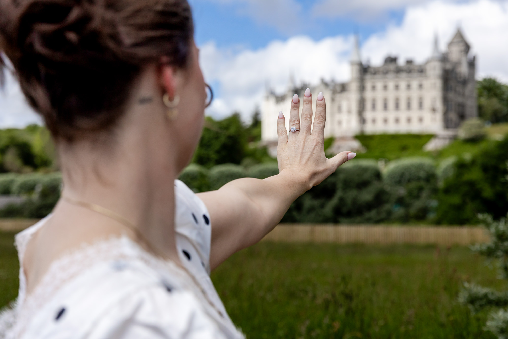 woman looking at her diamond engagement ring, with Dunrobin Castle in the background in the Scottish Highlands. 