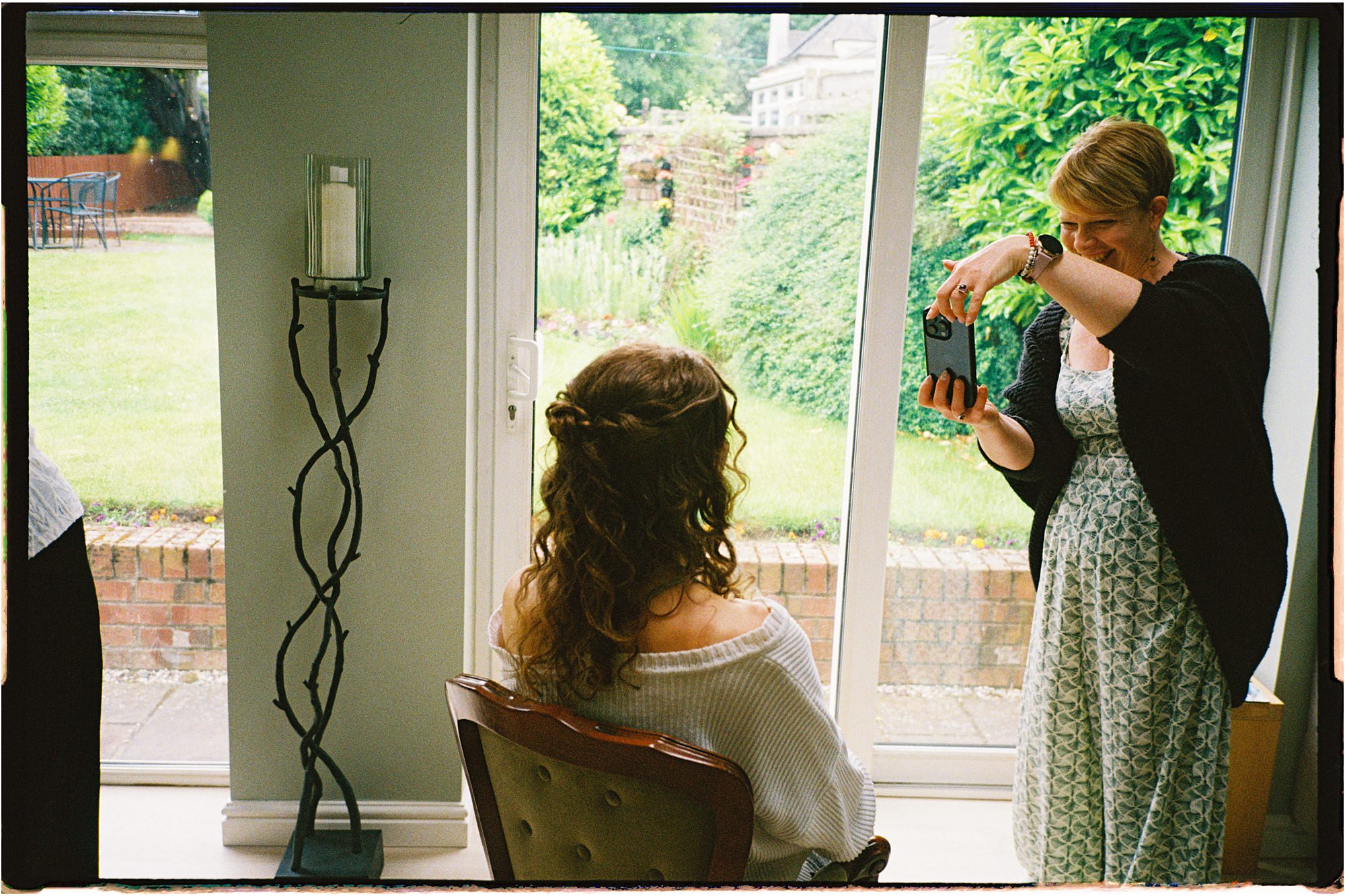 analogue photo of Rachel Spence HMUA taking a phone picture of a bride at home, in a bright room with large windows