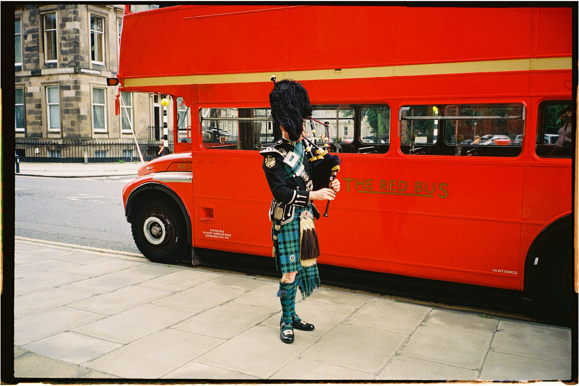 PIper Glyn Morris, playing the bagpipes in Highland dress, next to a red bus in Edinburgh taken on 35mm film