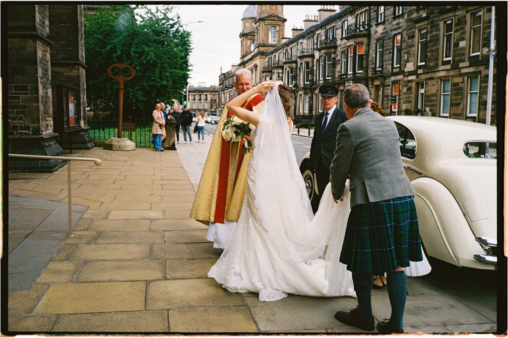 bride holding her veil as her parents attend her, standing next to a minister outside St Mary's Episcopal Church in Edinburgh, taken on 35mm film