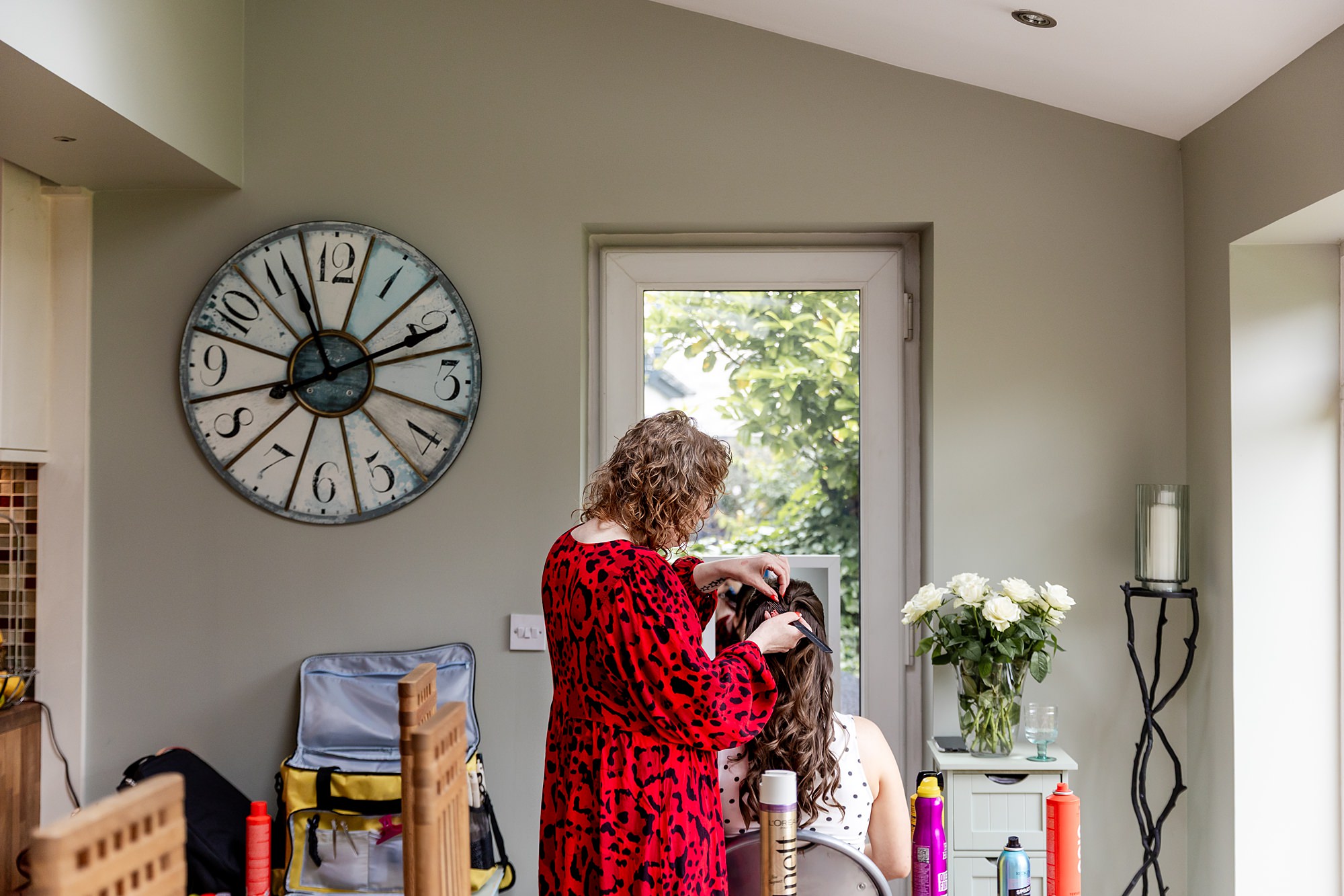 Hair stylist pinning a bridesmaids hair, at a home setting in a sun room at a Scottish house. There is a large analogue clock on the wall and white flowers in a vase.